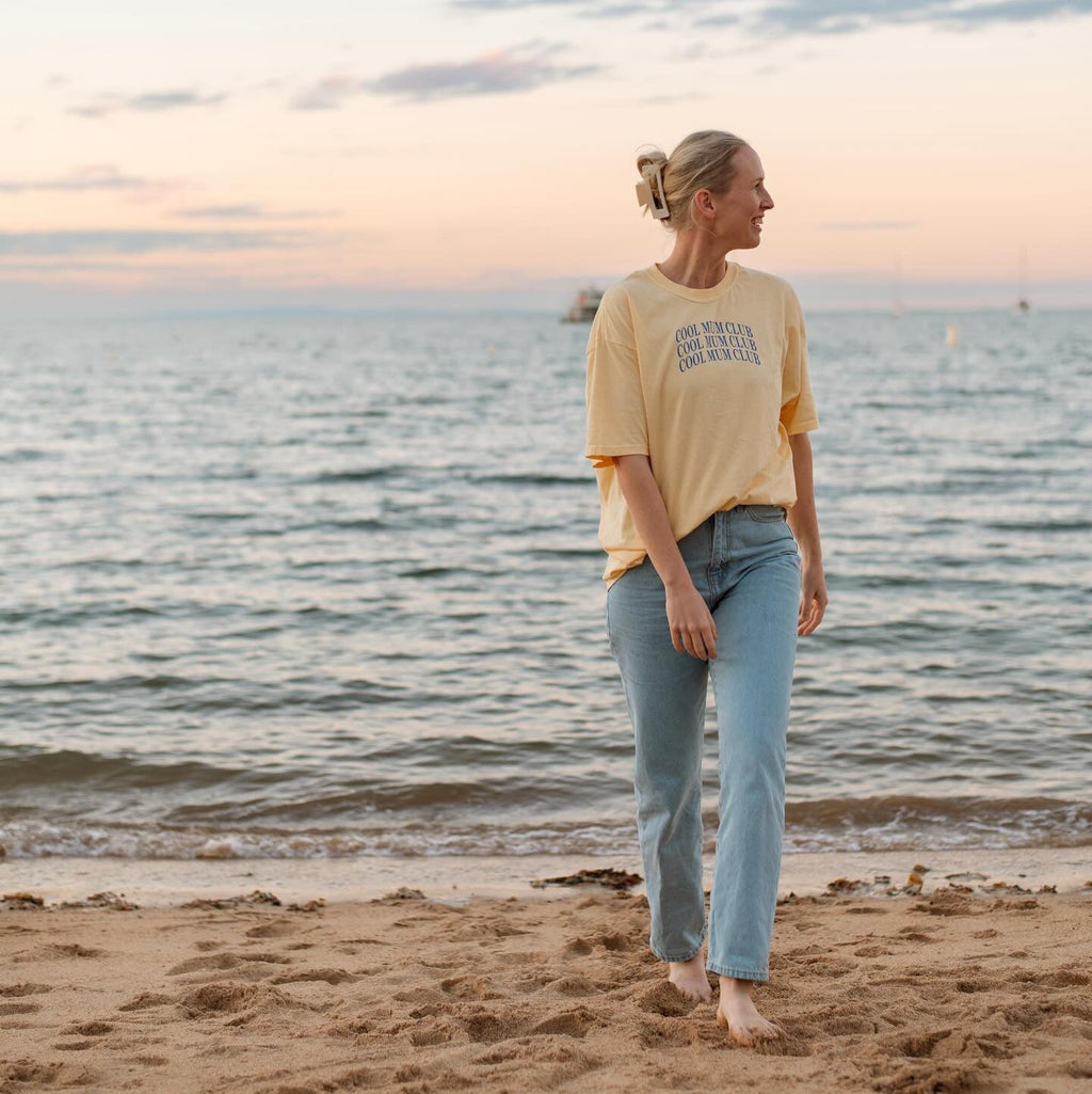 Woman walking on a beach at sunset wearing a yellow t-shirt and blue jeans.