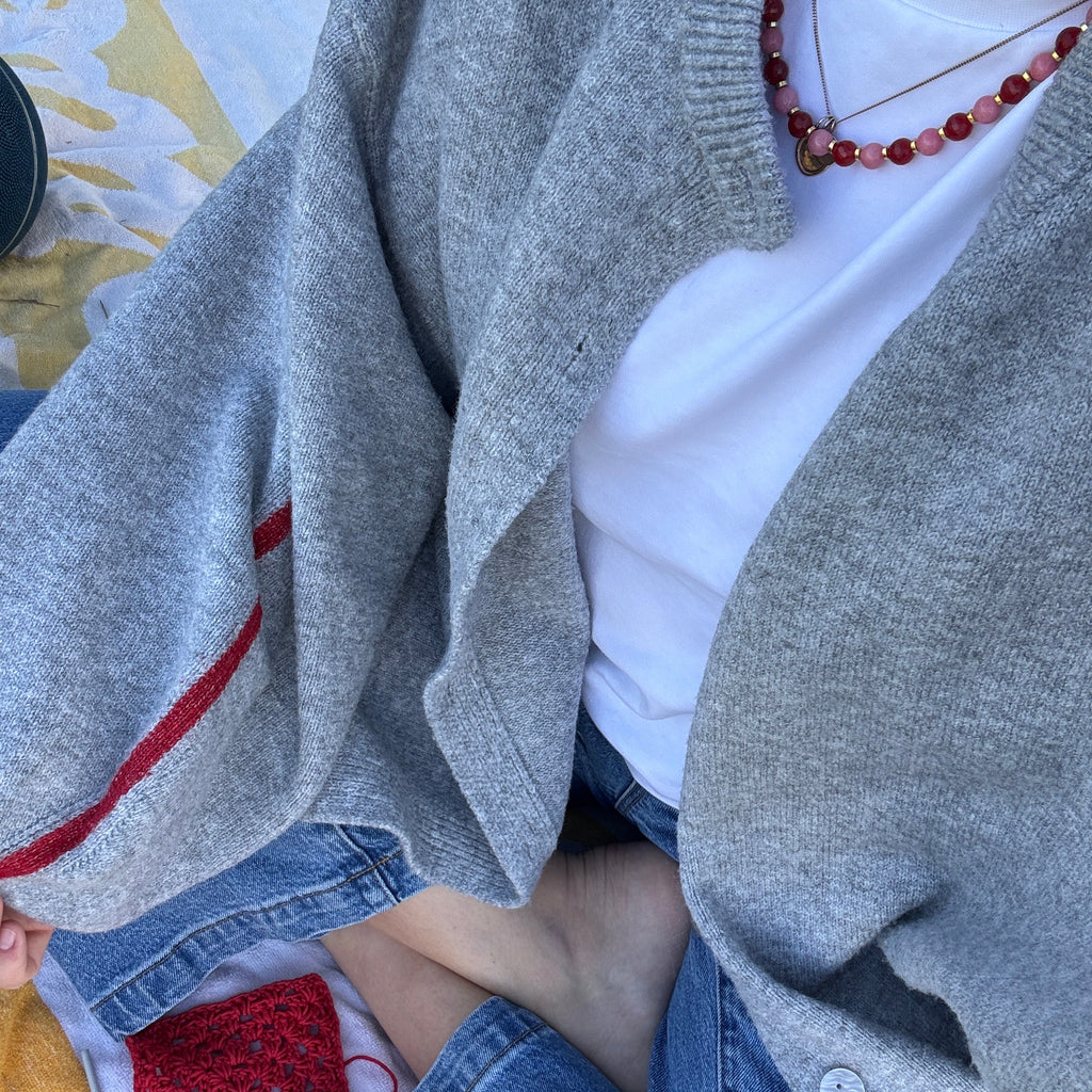 Person wearing a gray sweater and blue jeans sitting on a colorful blanket wearing a pink and red necklace.