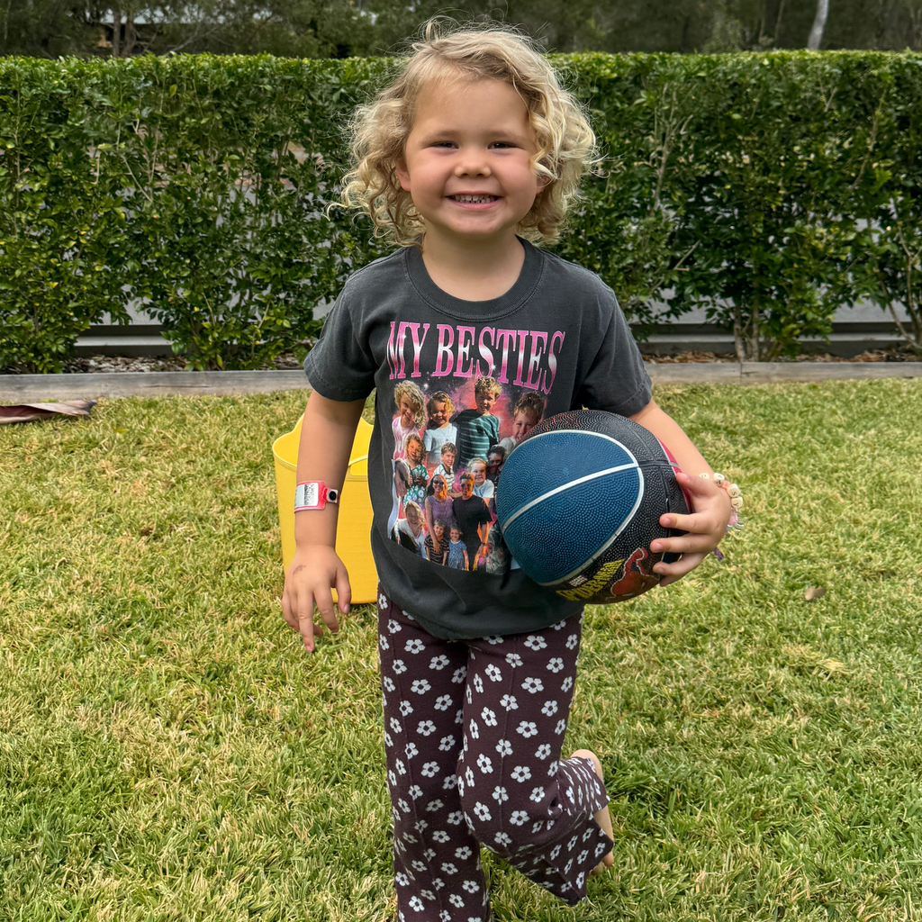 A young girl with curly blonde hair stands on a green lawn, smiling and holding a small blue basketball. She is wearing a dark gray graphic t-shirt with the text 'MY BESTIES' and dark patterned pants with white paw prints.