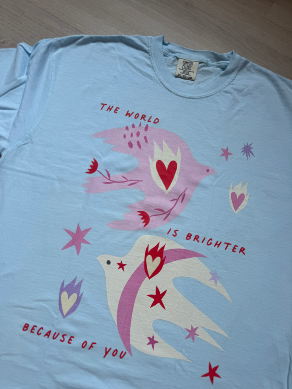 Light blue t-shirt with colorful bird design and text on a wooden surface