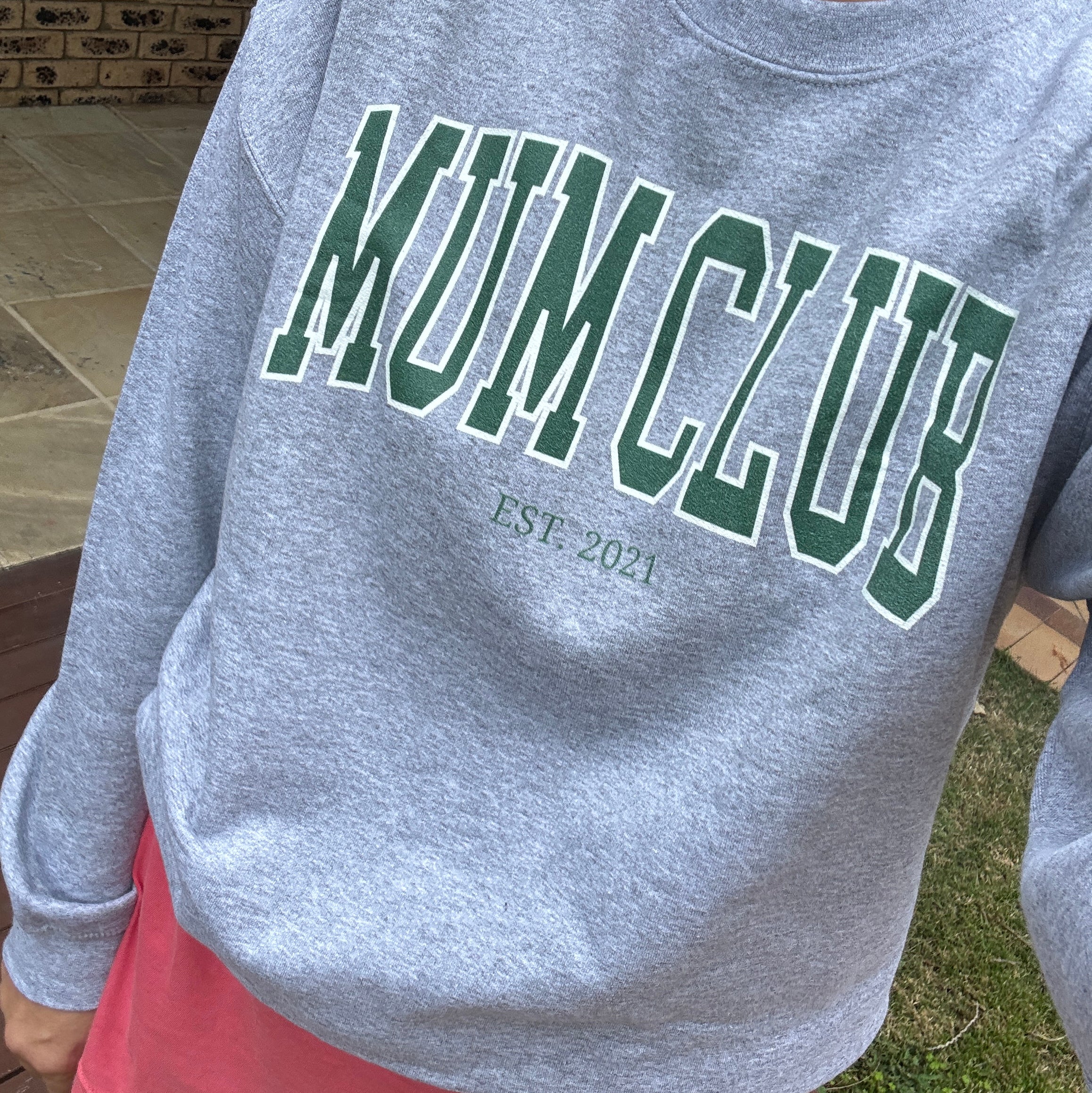 Gray sweatshirt with 'MUM CLUB EST 2021' text on a person wearing it outdoors.