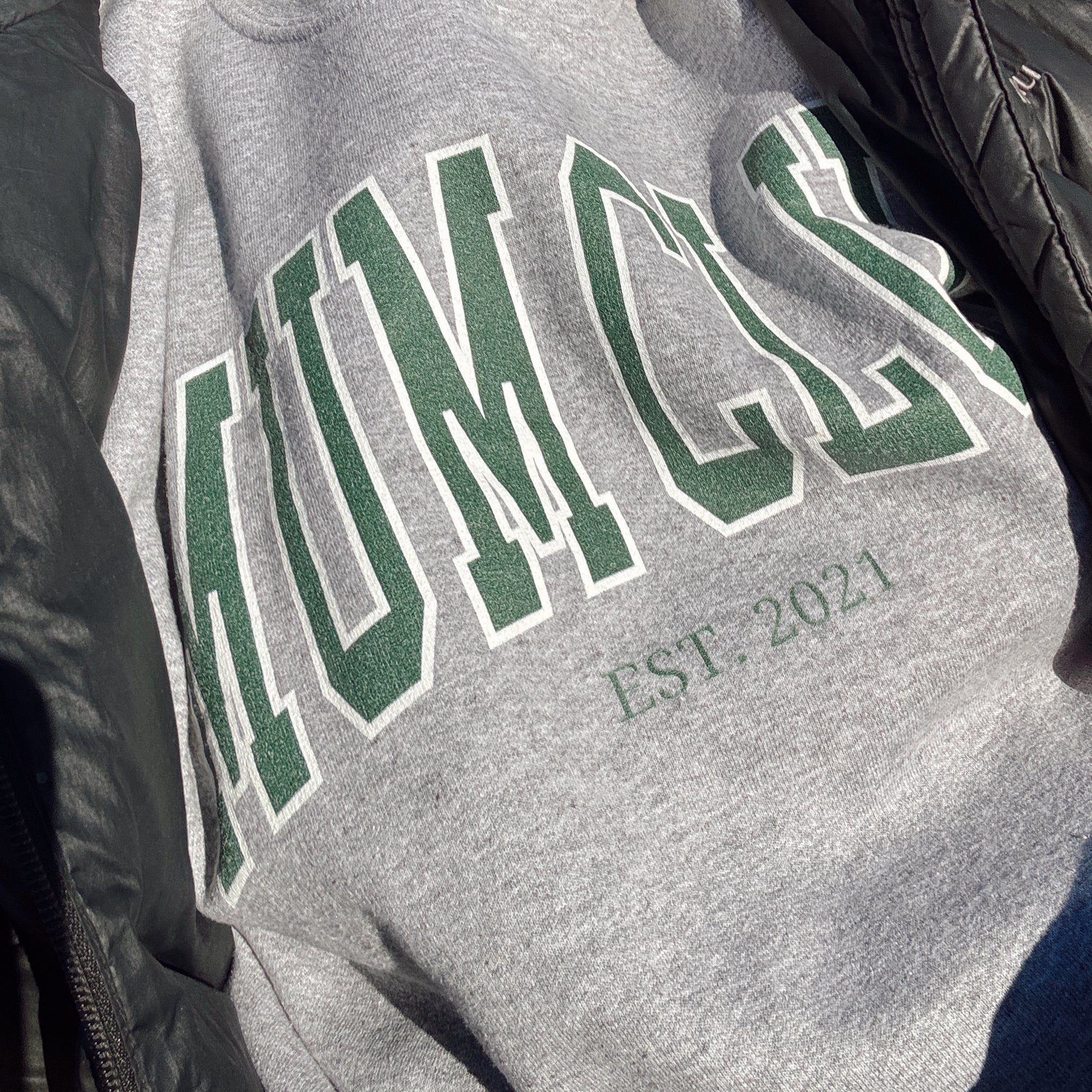 Gray sweatshirt with green text on a person wearing a black jacket.